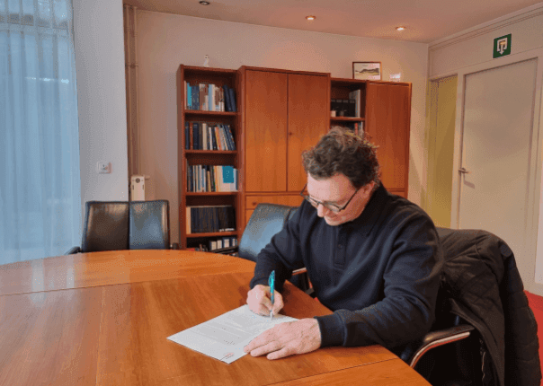 Man ondertekent een document aan een houten vergadertafel in een kantoor met boekenkasten