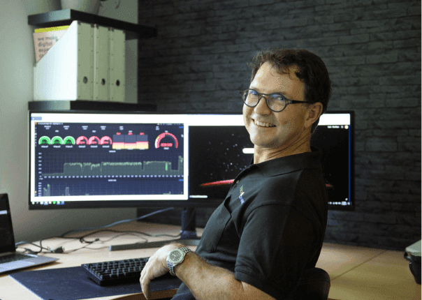 Lachende man met bril aan zijn bureau voor twee beeldschermen met een energiemonitoringdashboard met meters en verbruiksgrafieken